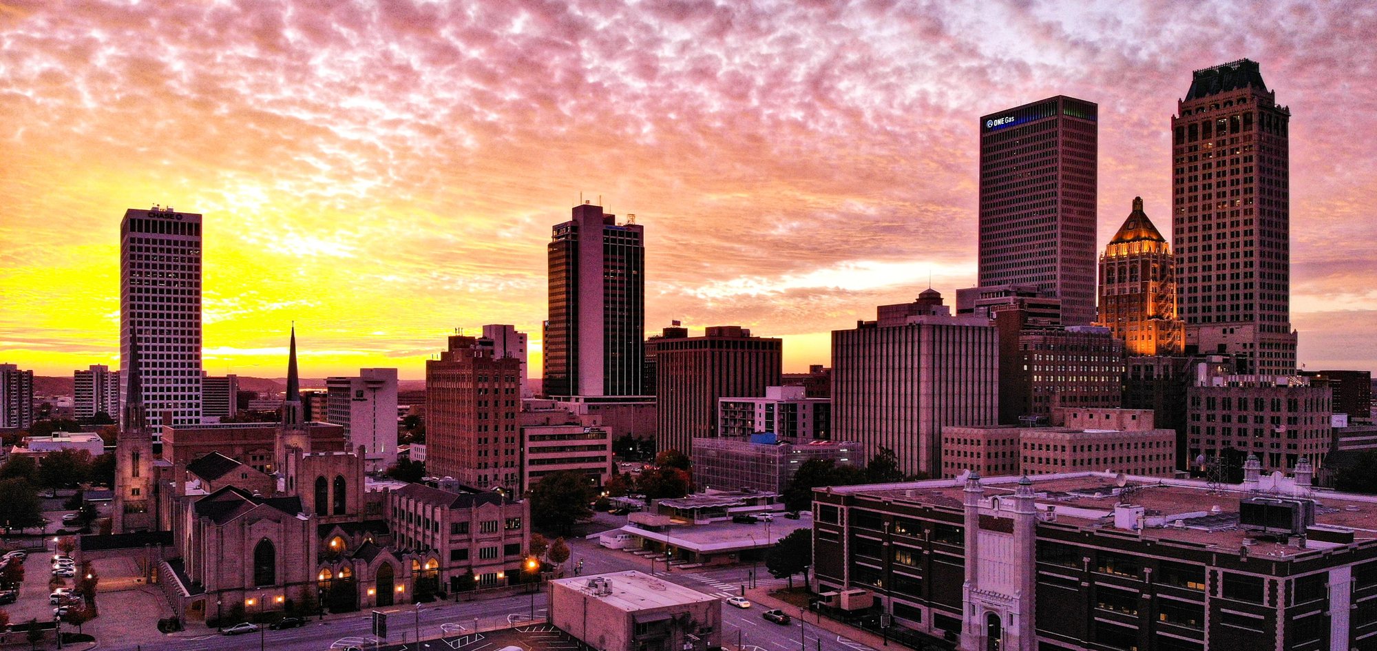 Downtown Tulsa skyline at sunrise, with office towers and a church spire under a pink and orange sky