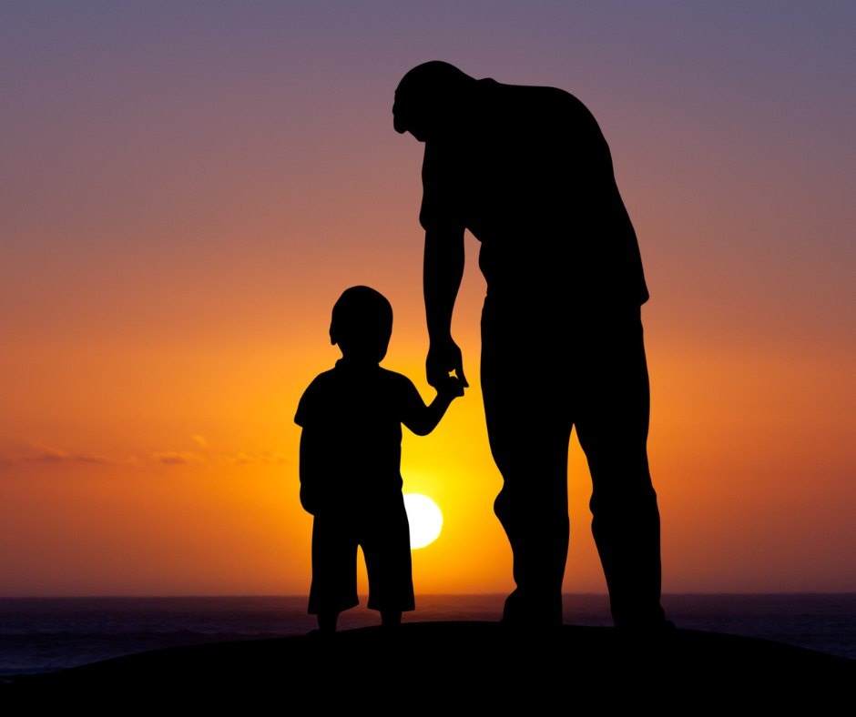 Silhouette of a father holding his young son’s hand as the sun sets over the water