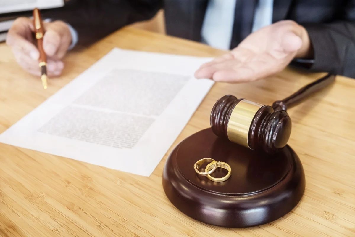 Two gold wedding rings on a gavel block beside a legal document on a wooden desk, with a person in a suit seated in the background