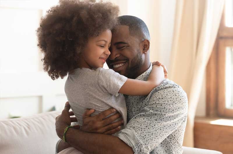 A smiling father hugging his young child on a sofa near a sunlit window